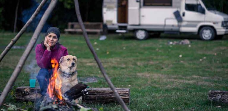 Frau sitzt mit Hund am Lagerfeuer auf einem Campingplatz